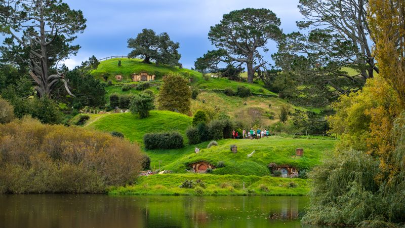 Round hobbit-hole doors built into grassy hillsides above a still pond, framed by tall trees