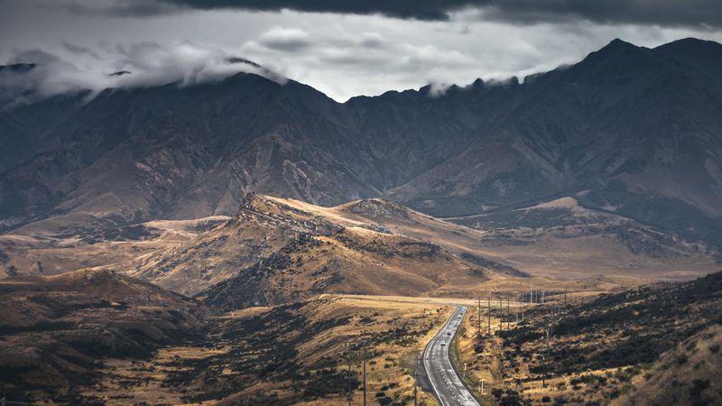 Two-lane highway winding through a rugged mountain valley beneath a stormy sky