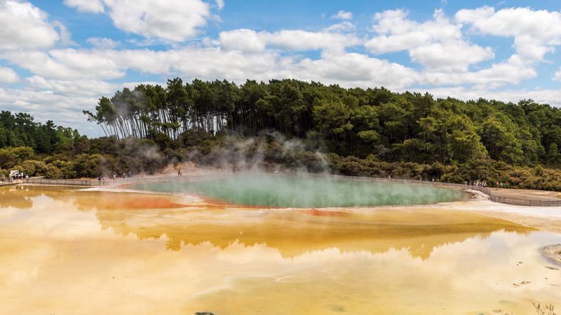 Steaming, brightly colored geothermal pool bordered by forest under a partly cloudy sky